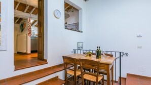 Un coin repas avec une table et des chaises en bois, une horloge au mur et une fenêtre donnant sur une cuisine.