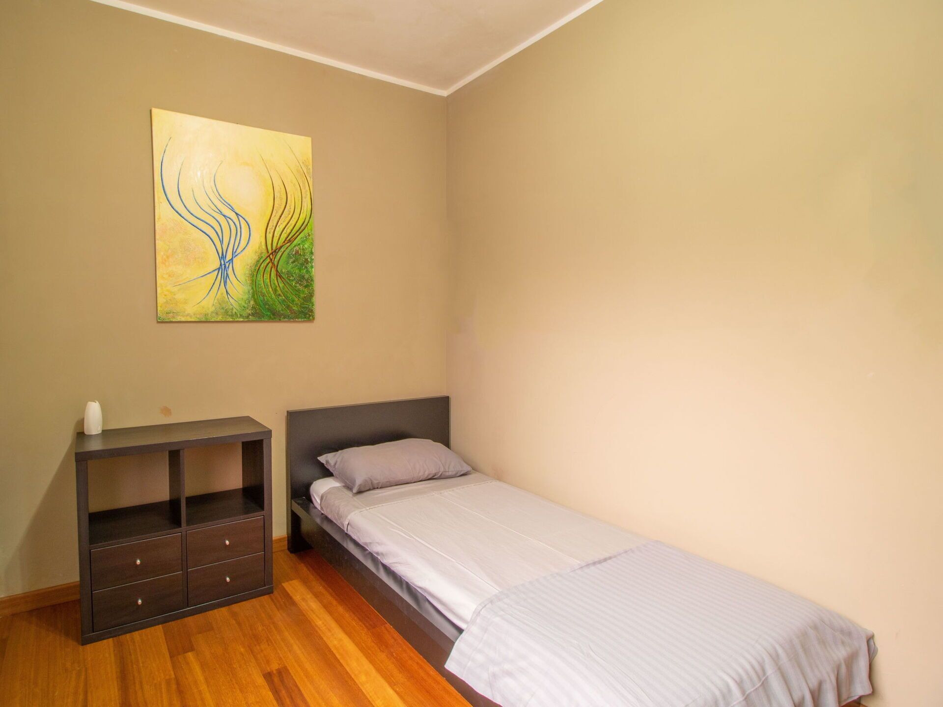 A bedroom with a single bed, a nightstand, a dresser, and a wall-mounted abstract painting.