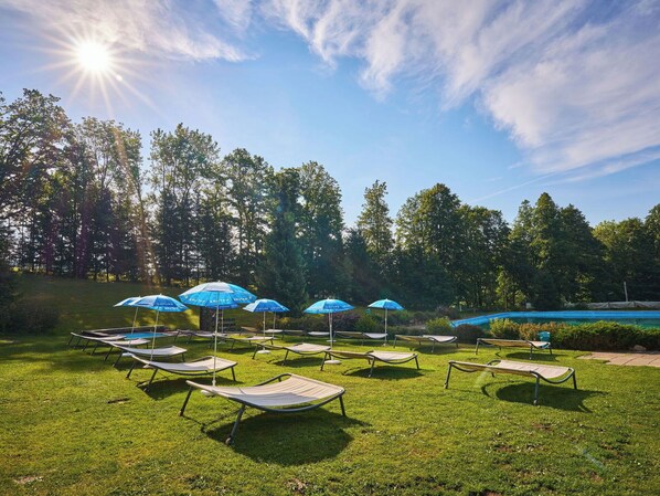 A grassy area with sun loungers and blue umbrellas, surrounded by trees.