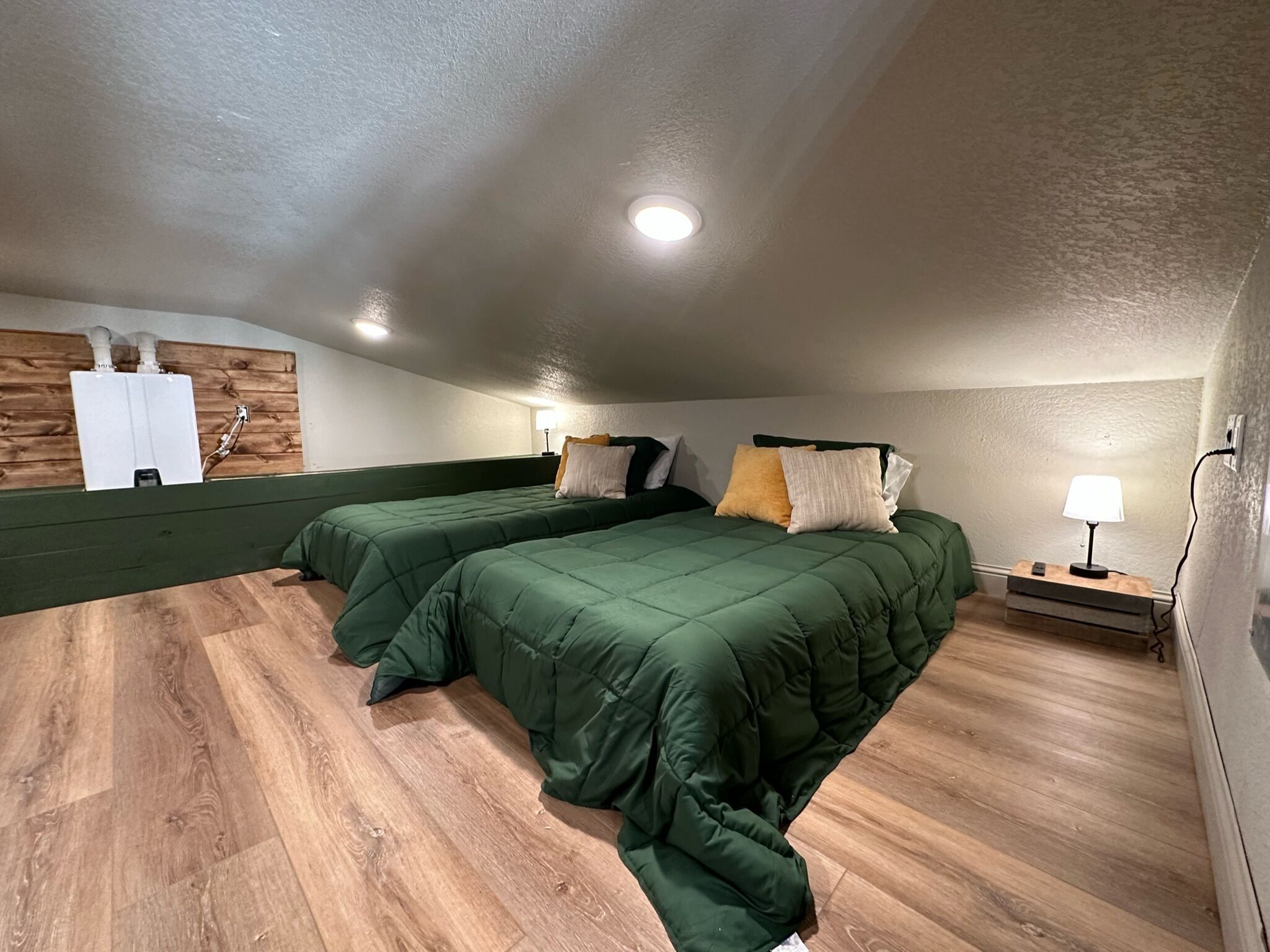 Quarto com duas camas, piso de madeira, aquecedor fixado na parede e mesa de cabeceira com abajur.