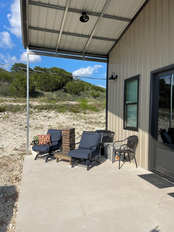 Ein überdachter Patio-Bereich mit Sitzgelegenheiten im Freien, darunter zwei Liegestühle und ein kleiner Tisch, mit Blick auf eine natürliche Landschaft.