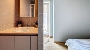 Ein modernes Badezimmer mit einem großen Spiegel, einem Waschbecken und zwei Blumentöpfen. Der Boden und die Wände sind in einem hellen Farbton gehalten und es gibt eine Tür zu einem weiteren Raum.