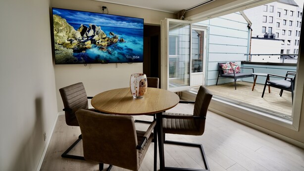 A modern living room with a round wooden table, four brown chairs, a large flat-screen TV on the wall, and a balcony with a view of buildings.