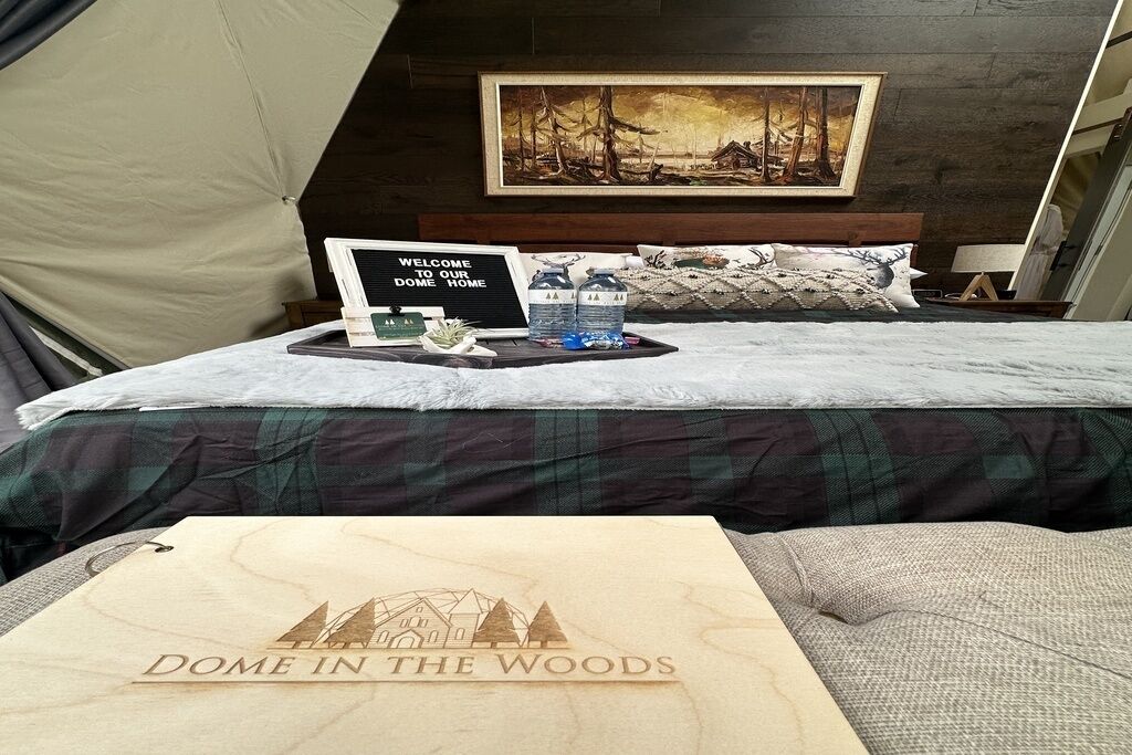 Une pièce chaleureuse avec un lit, une pancarte de bienvenue, un tableau et une enseigne en bois sur laquelle on peut lire : « DOME IN THE WOODS ».