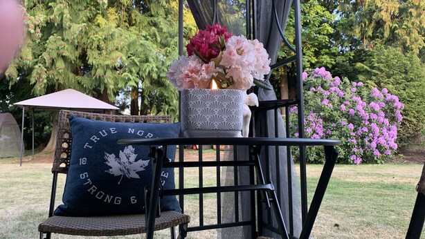 Un espace extérieur abrité, aménagé avec des chaises en osier, un banc rembourré et une table agrémentée d’un vase de fleurs.