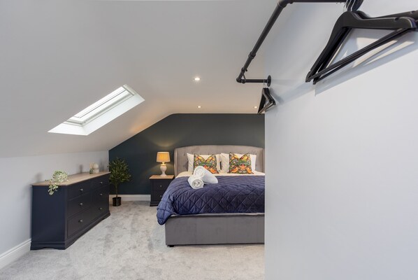 A bedroom with a sloped ceiling, a skylight, a bed with a dark blue quilt, two decorative pillows, a nightstand with a lamp, and a dresser with drawers. - Kist Accommodates - Belmont Dene (North Yorkshire Hub) (Knaresborough)