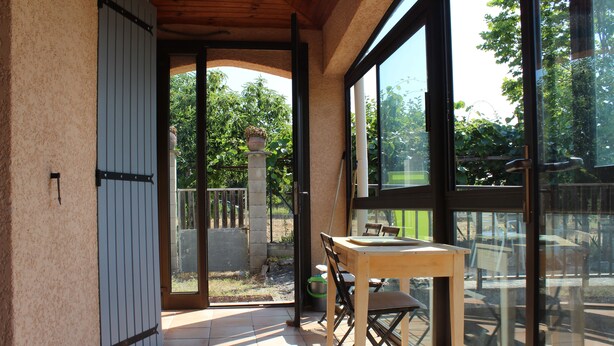 Eine Terrasse mit Tisch und Stühlen, eine Glasschiebetür, die zu einem Garten führt, und Blick auf Bäume und einen Zaun.