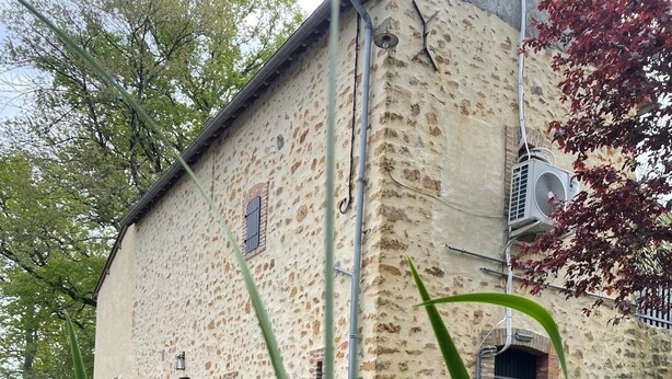 Un bâtiment rustique en pierre, doté d’une petite fenêtre et d’une porte, entouré de verdure et agrémenté d’un élément décoratif aquatique.