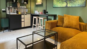 Un espace de vie compact comprenant un coin cuisine, une table basse en verre, un canapé de couleur moutarde et un tableau représentant un arbre.
