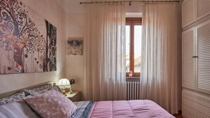 A bedroom with a bed, a nightstand, a window with curtains, and a radiator.