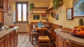 A cozy kitchen with wooden cabinets, a dining table, and a bench. There are decorative plates on shelves, a window with curtains, and a fruit bowl on the counter.