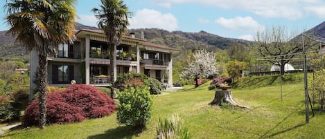 A two-story building with balconies, surrounded by a well-maintained garden with various shrubs and a palm tree.