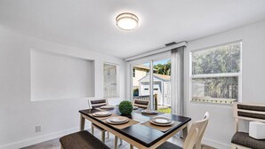 Uma sala de jantar com uma mesa posta para quatro pessoas, cadeiras, uma planta pequena e vista para o exterior.