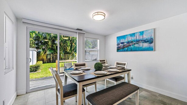 Uma sala de jantar com mesa posta para seis pessoas, um banco e vista para um jardim tropical através de portas de vidro.