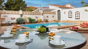 Une table en verre avec des tasses blanches et des pâtisseries étiquetées « Paradise Sweet », une piscine et un bâtiment blanc doté de fenêtres en arc de cercle.