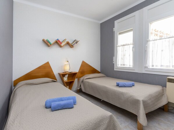 A hotel room with two beds, a bedside table with a lamp, a window with curtains, and a wall-mounted bookshelf.