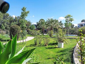 Um jardim com um caminho, diversas plantas, uma piscina e um edifício ao fundo.