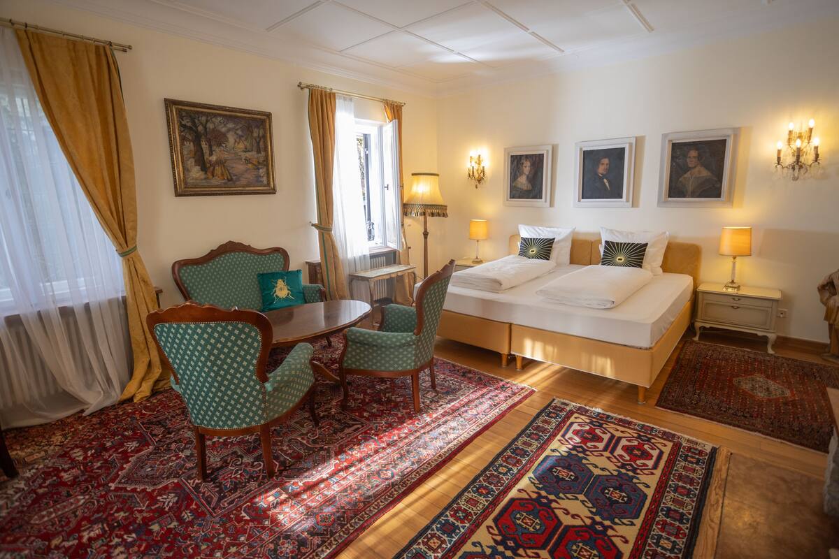A hotel room with a bed, two chairs, a table, a lamp, and a painting on the wall.