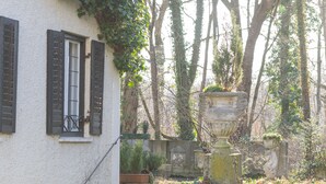 Ein weißes Gebäude mit schwarzen Fensterläden, einer Steintreppe und einem großen Urnenbrunnen im Garten.