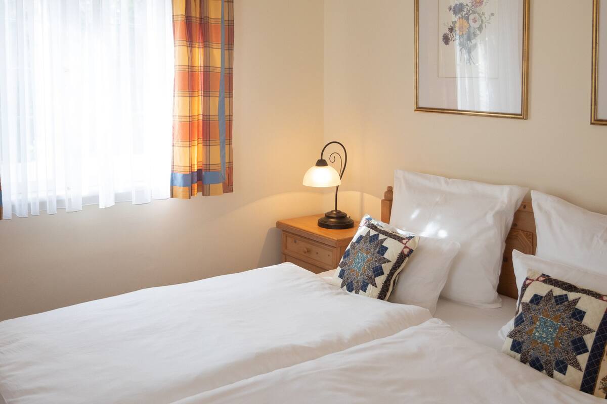 A bedroom with a large bed, a nightstand with a lamp, a window with curtains, and a chandelier.