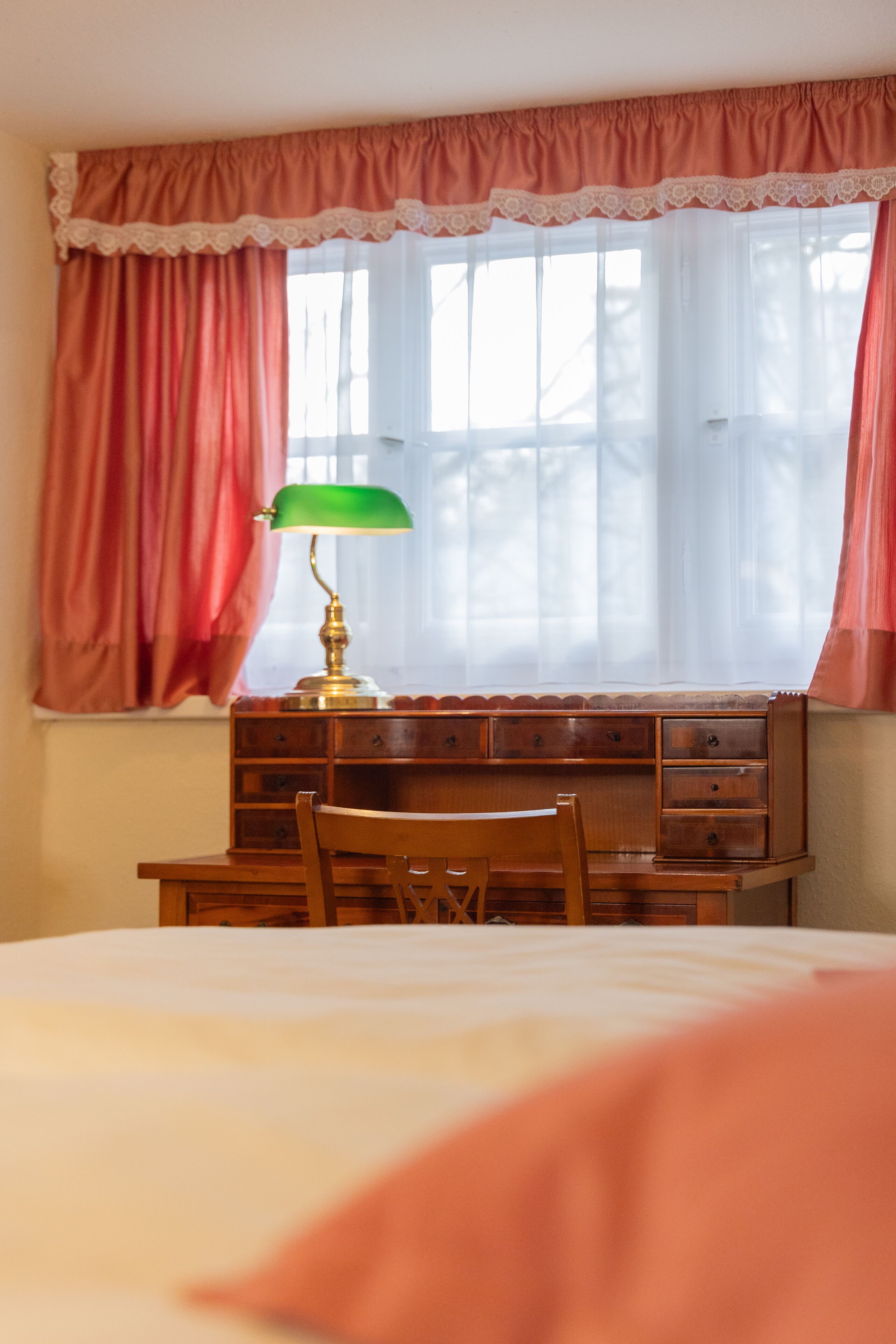 A bedroom with a bed, a wooden dresser, a desk with a lamp, and a window with curtains.