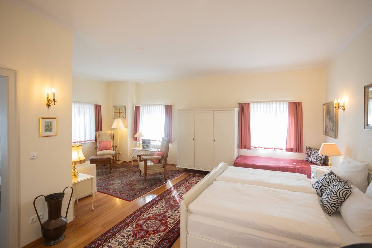 A hotel room with a large bed, a desk with a lamp, a chair, a wardrobe, and a red carpet.