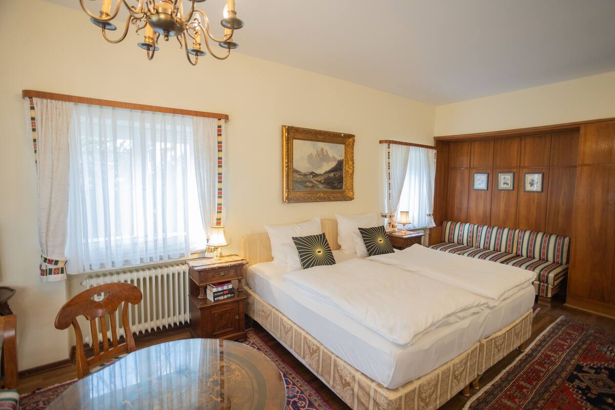 A well-lit bedroom with a large bed, a wooden wardrobe, a striped sofa, a round table, and a chandelier.