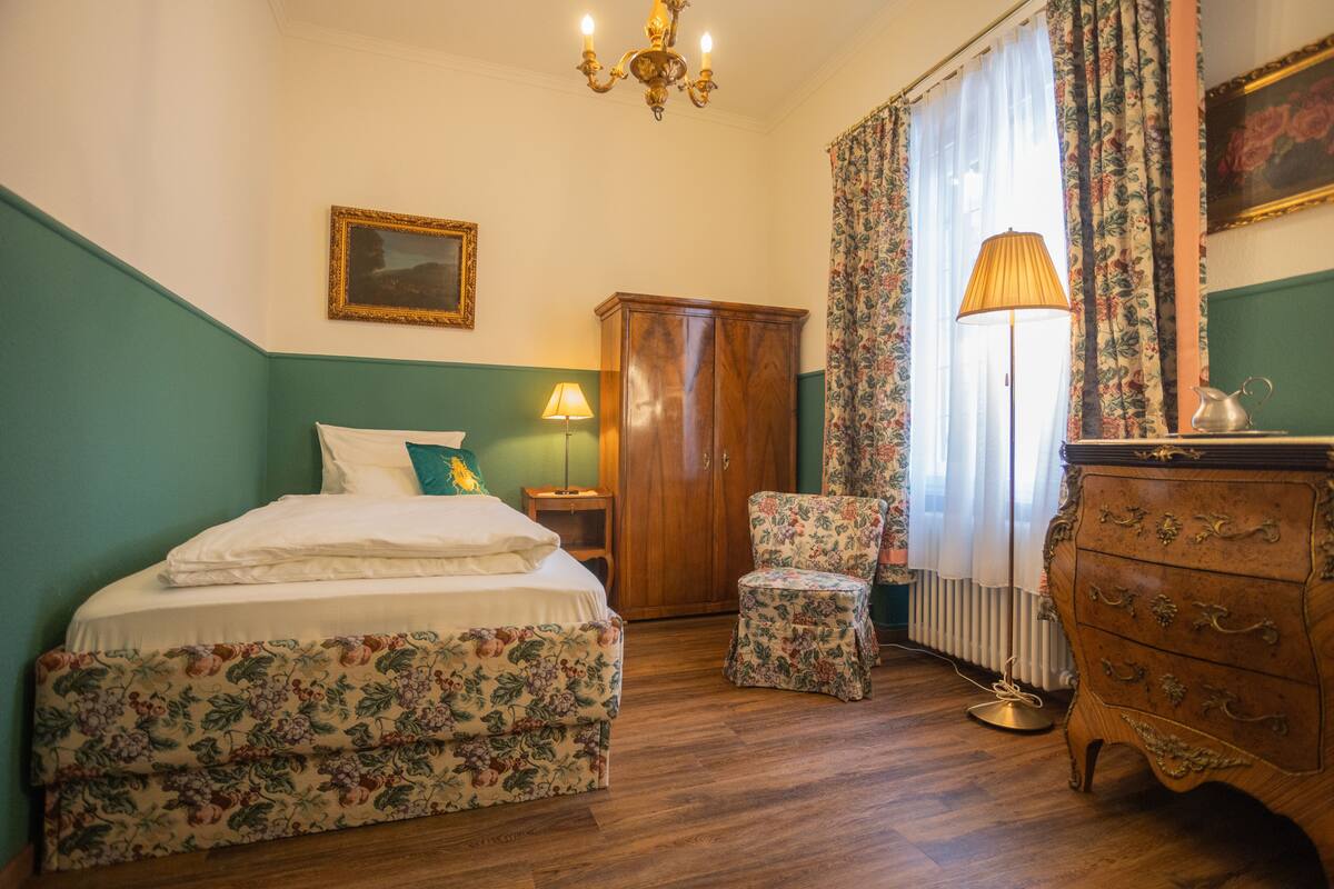 A bedroom with a bed, a wooden wardrobe, a chair, a lamp, and a radiator.