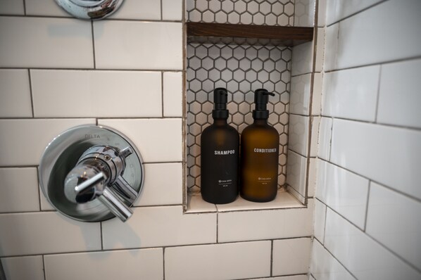 Um banheiro com parede azulejada, chuveiro e dois frascos de shampoo e condicionador em uma prateleira.