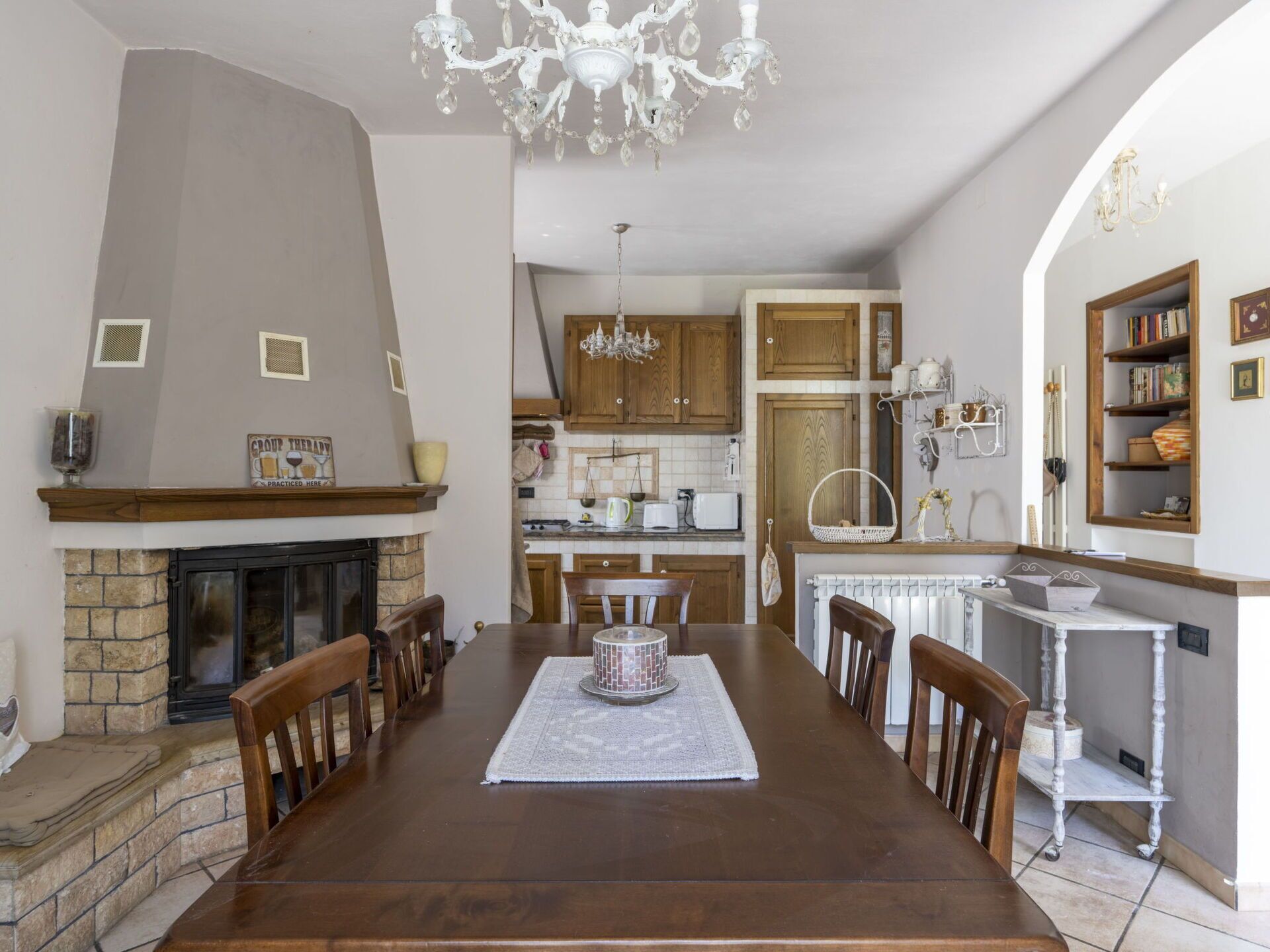 Un comedor con chimenea, sillas de madera, una araña y una cocina al fondo.