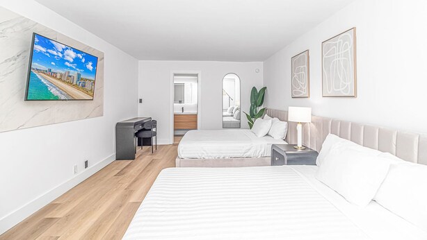 A modern hotel room with two beds, a desk, a mirror, and a TV on the wall. - Queen Suite/Fifth and Ocean (Myrtle  Beach)