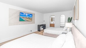 A modern hotel room with a large TV mounted on a marble wall, a bed with white linens, a desk with a chair, a mirror, and a plant. - Queen Suite/Fifth and Ocean (Myrtle  Beach)