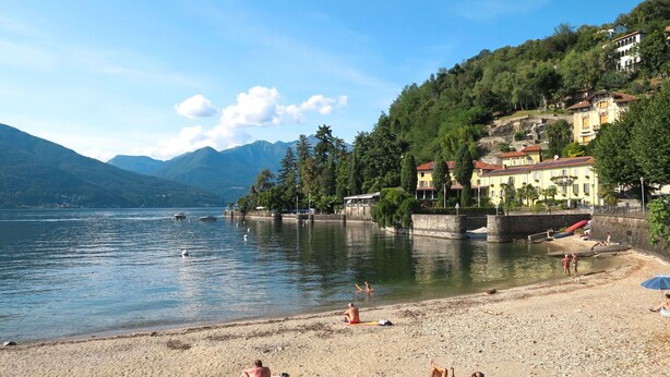 Ein Strand am See mit badenden Personen, sandigem Ufer und Gebäuden im Hintergrund.
