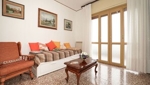 Ein Wohnzimmer mit einer Couch, einem Sessel und einem Couchtisch. An den Wänden hängen zwei Bilder und ein großes Fenster mit Vorhängen.