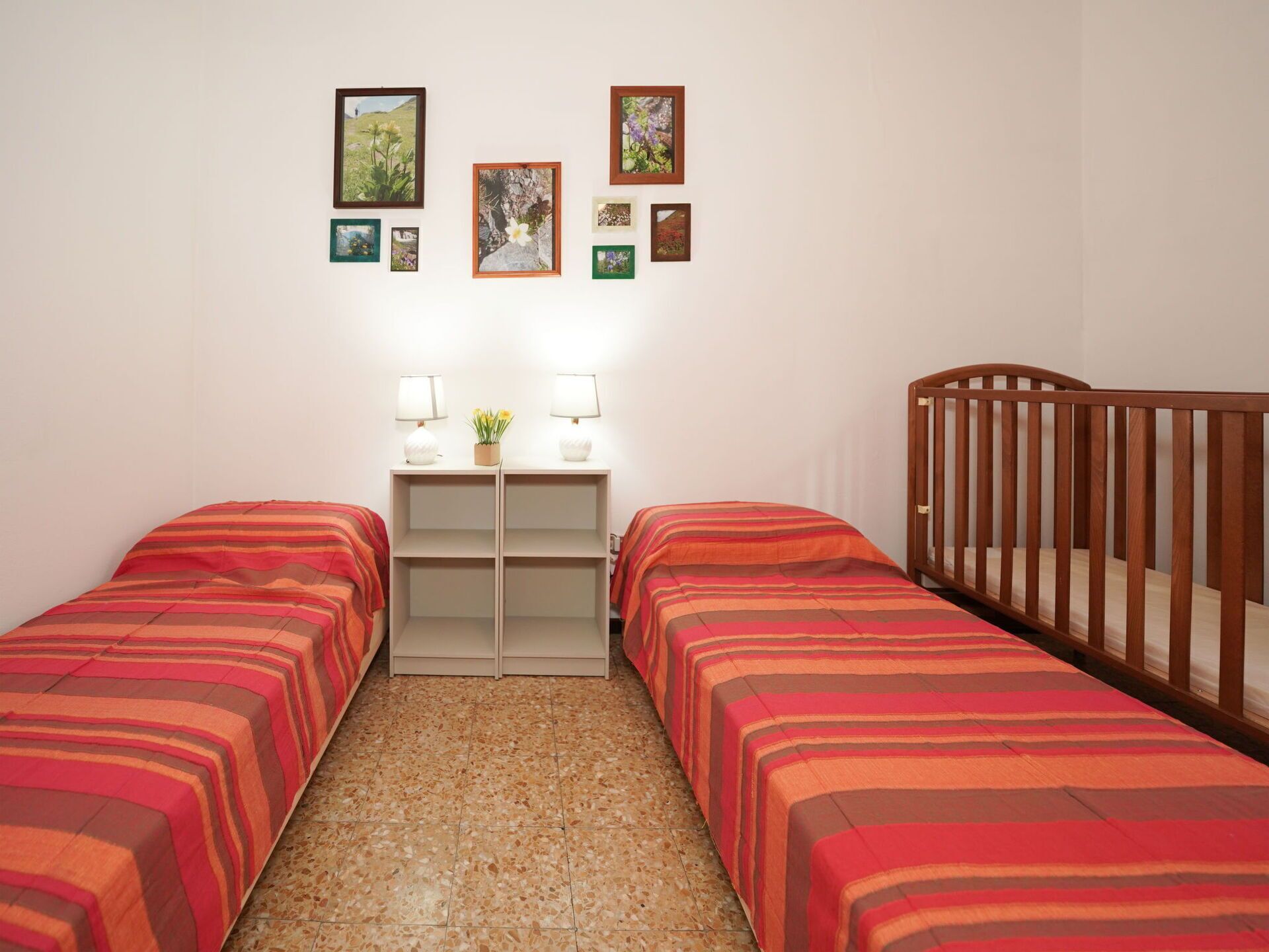 Ein Zimmer mit zwei Einzelbetten, einem Holz-Babybett, einem kleinen Schreibtisch mit zwei Lampen und einer Wand mit gerahmten Bildern.