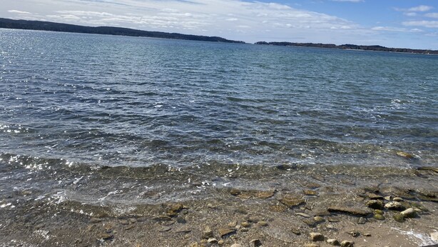 A clear view of a lake with a rocky shoreline and distant land. - M-22 Lakefront Property in Onekama with amazing Sunsets!! (Onekama)