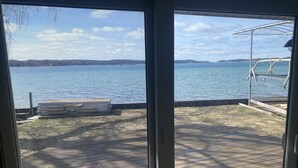 A view from inside a room with a large window overlooking a lake, a dock with a boat, and a wooden deck. - M-22 Lakefront Property in Onekama with amazing Sunsets!! (Onekama)