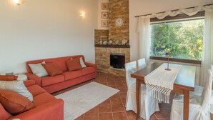 A living room with a red sofa, a fireplace, a dining table with chairs, and a view of greenery outside.