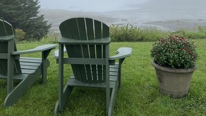 Two Adirondack chairs on a grassy area with a view of the ocean and a foggy backdrop.