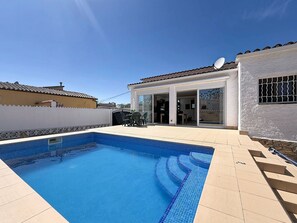 Une piscine avec des marches carrelées, une terrasse avec des chaises et une maison équipée d’une antenne parabolique.