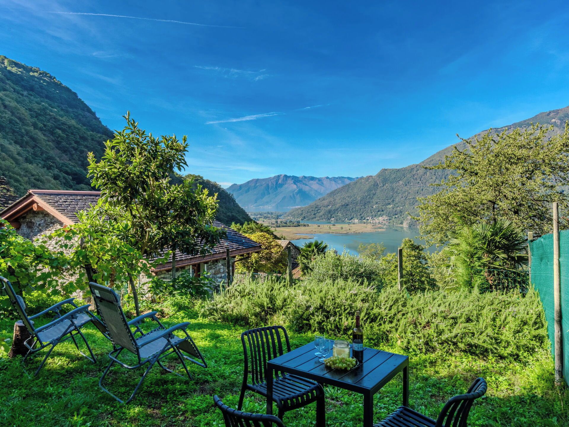 Una mesa con sillas y una botella, rodeada de follaje y con vistas a montañas y un lago.