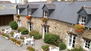 A stone building with multiple windows and a covered outdoor seating area with white chairs and planters with red flowers. - Nice apartment for 5 guests with WIFI, pool, TV and terrace (Saint-jouan-des-guérets)