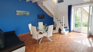 A dining area with a glass table and white chairs, a blue wall, a staircase, and a view of greenery outside. - Nice apartment for 5 guests with WIFI, pool, TV and terrace (Saint-jouan-des-guérets)