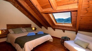 Un dormitorio con techo de madera, una cama con manta verde, una mesita de noche con lámpara y vistas a las montañas a través de un tragaluz.
