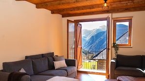 Una sala de estar acogedora con techo de madera, un sofá gris, una mesita de centro y vistas a las montañas a través de puertas abiertas.