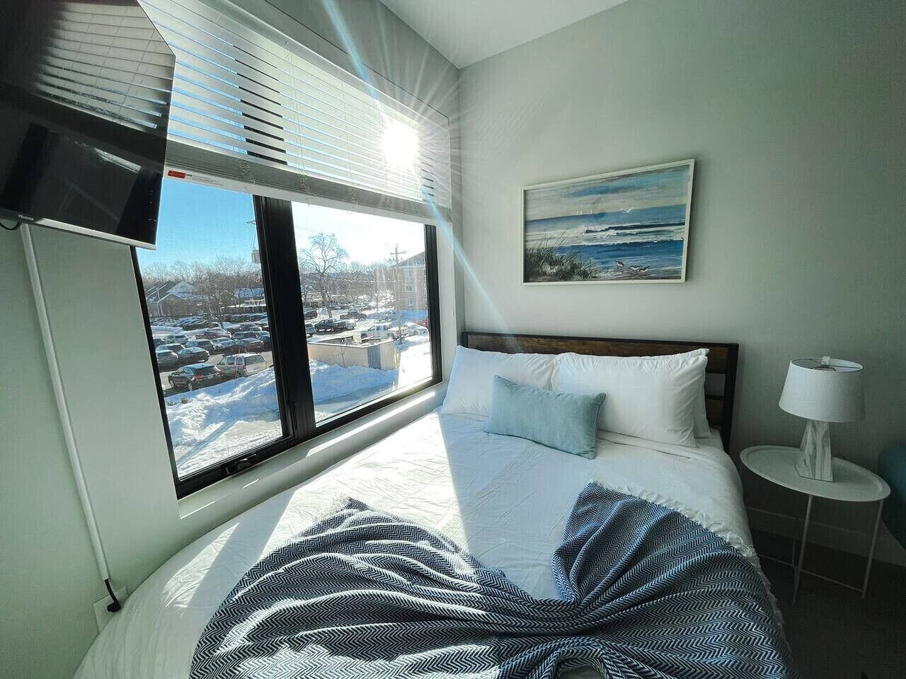 A bedroom with a large window, a bed with white and blue bedding, a painting on the wall, and a bedside lamp.