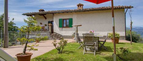 A rustic house with a stone facade, green shutters, and a patio with outdoor furniture and potted plants.