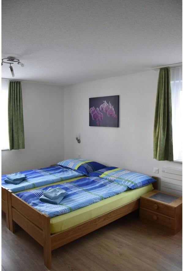 A single-bed room with a wooden frame, a green curtain, a small bedside table, and a wall-mounted light fixture.