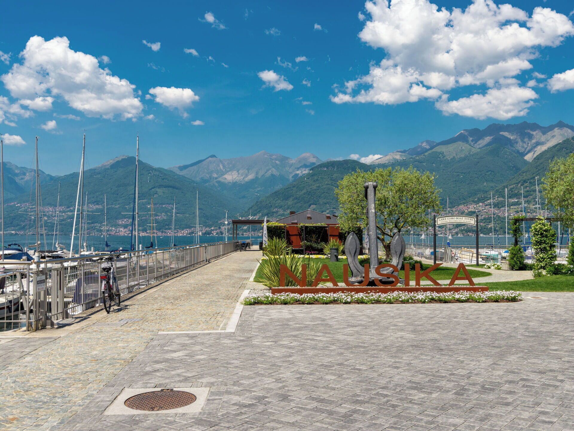 A waterfront promenade with a sign for 'NAUSIKA' and a view of boats and mountains in the background.