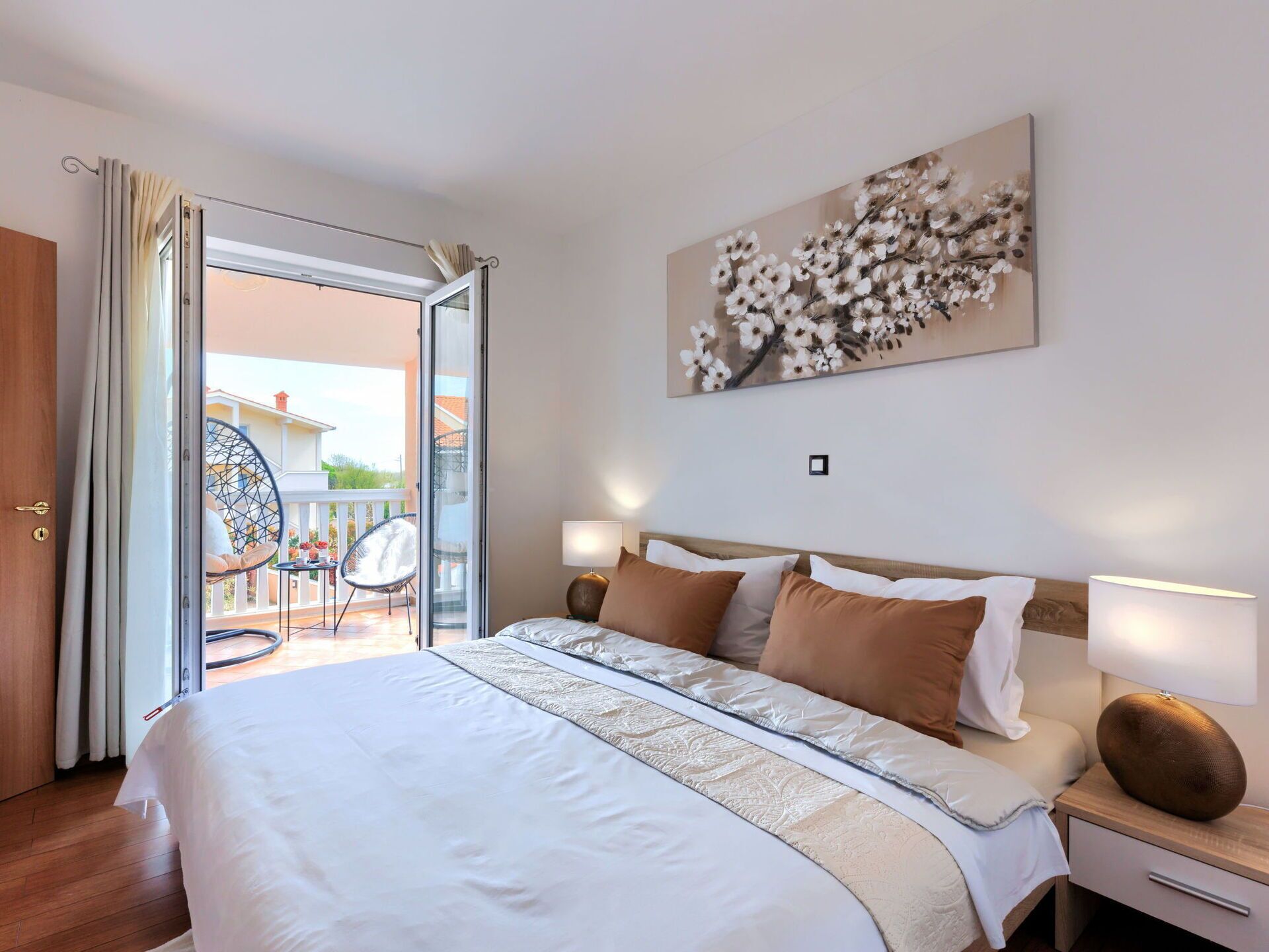 A hotel room with a bed, bedside tables, a lamp, a painting, and an open door leading to a balcony with a chair and table.